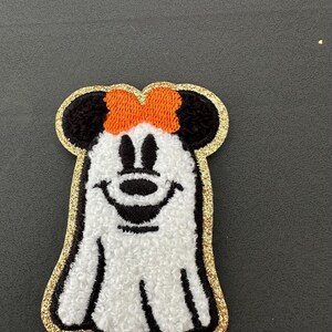 May include: A white ghost with a black outline and a black and white face. The ghost has a large orange bow on its head. The ghost is outlined in gold glitter.