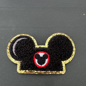 May include: A black and gold Mickey Mouse ear hat patch with a red and white Mickey Mouse head in the centre.