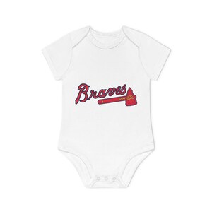 Può includere: Body da bambino bianco con la scritta "Braves" in corsivo rosso e un logo a forma di tomahawk rosso e oro. Il body ha maniche corte e chiusura con bottoni a pressione.