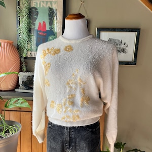 May include: A white sweater with a delicate floral pattern and gold bead embellishments. The sweater has long sleeves and a crew neckline.