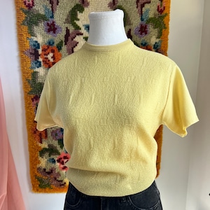 May include: A pale yellow short-sleeved top with a textured pattern. The top is displayed on a mannequin, paired with black denim jeans. The background features a colorful tapestry and a white decorative screen.