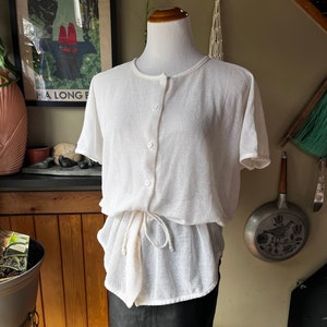 May include: White mesh button-up top with short sleeves and a drawstring waist, paired with a black leather skirt.