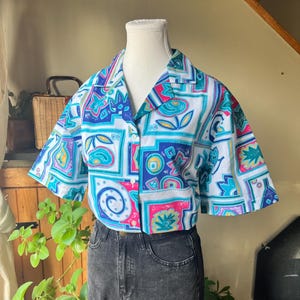 May include: A short-sleeved shirt with a colourful geometric design in blue, pink, and teal on a white base. The shirt is worn with dark grey denim jeans. The shirt is displayed on a mannequin.