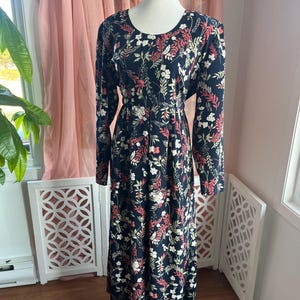 May include: A long, black dress with a floral pattern of white and red flowers and green leaves. The dress has long sleeves and a scoop neckline. The dress is displayed on a mannequin.