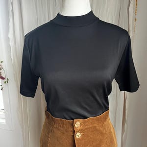 Puede incluir: Una camiseta negra de manga corta con cuello alto combinada con una falda de pana marrón. La falda tiene grandes botones dorados. La ropa se exhibe en un maniquí.