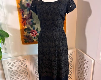 Puede incluir: Vestido negro vintage con una superposición de encaje floral. El vestido presenta un escote redondo con detalles recortados, mangas cortas y una cintura entallada. La falda tiene un dobladillo negro liso. El vestido se exhibe en un maniquí blanco.