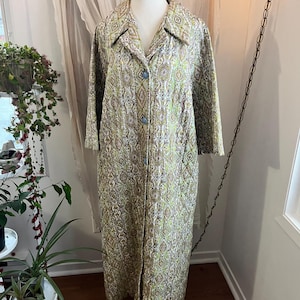 May include: A vintage, floor-length robe with a paisley pattern in shades of green, brown, and white. The robe has a collared neckline, three-quarter sleeves, and light blue buttons. It is displayed on a mannequin.
