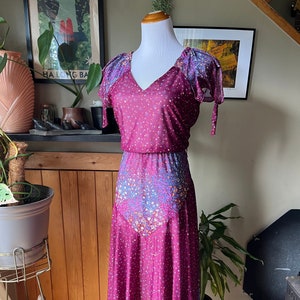 Puede incluir: Un vestido midi de estilo vintage con estampado floral en tonos morados, azules y blancos. El vestido tiene un escote en V, mangas cortas con lazos y un corpiño ajustado. La falda es fluida y cae hasta la mitad de la pantorrilla.