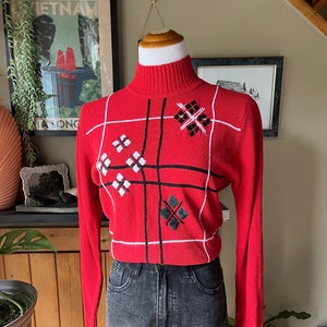 May include: Red turtleneck sweater with a black and white geometric pattern. The sweater has a mock neck and long sleeves.