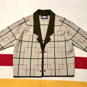 May include: A beige knit cardigan featuring a dark olive green lapel collar and trim. The garment displays a geometric grid pattern in black and thin red lines, with two gold-toned buttons at the center front and an interior label reading DEVALIA.