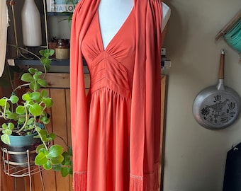 May include: A long, coral-colored dress with a V-neckline and a matching scarf. The dress has a fitted bodice and a flowing skirt. The scarf has fringe detailing.