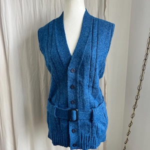 Puede incluir: Un chaleco de punto azul vintage con cuello en V, cierre de botones y cinturón con hebilla negra. El chaleco tiene dos bolsillos delanteros y detalles acanalados verticales. La prenda se exhibe en un maniquí blanco.