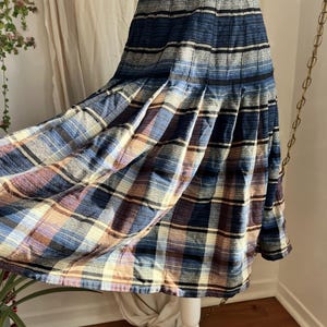 Op de afbeelding: Een vintage geruite rok in blauwe, paarse en crèmekleurige tinten. De rok heeft een geplooid ontwerp en een middellange zoom. De stof heeft een zacht, getextureerd uiterlijk, wat wijst op wol of een wolmix. De rok wordt gepresenteerd op een witte paspop.