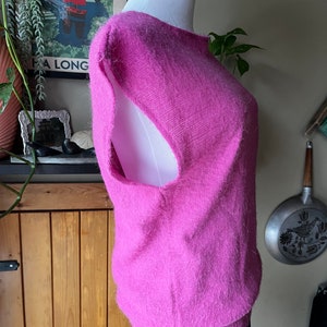 May include: A pink fluffy sleeveless sweater with a scoop neck. The sweater is on a white mannequin and is displayed against a wooden wall.