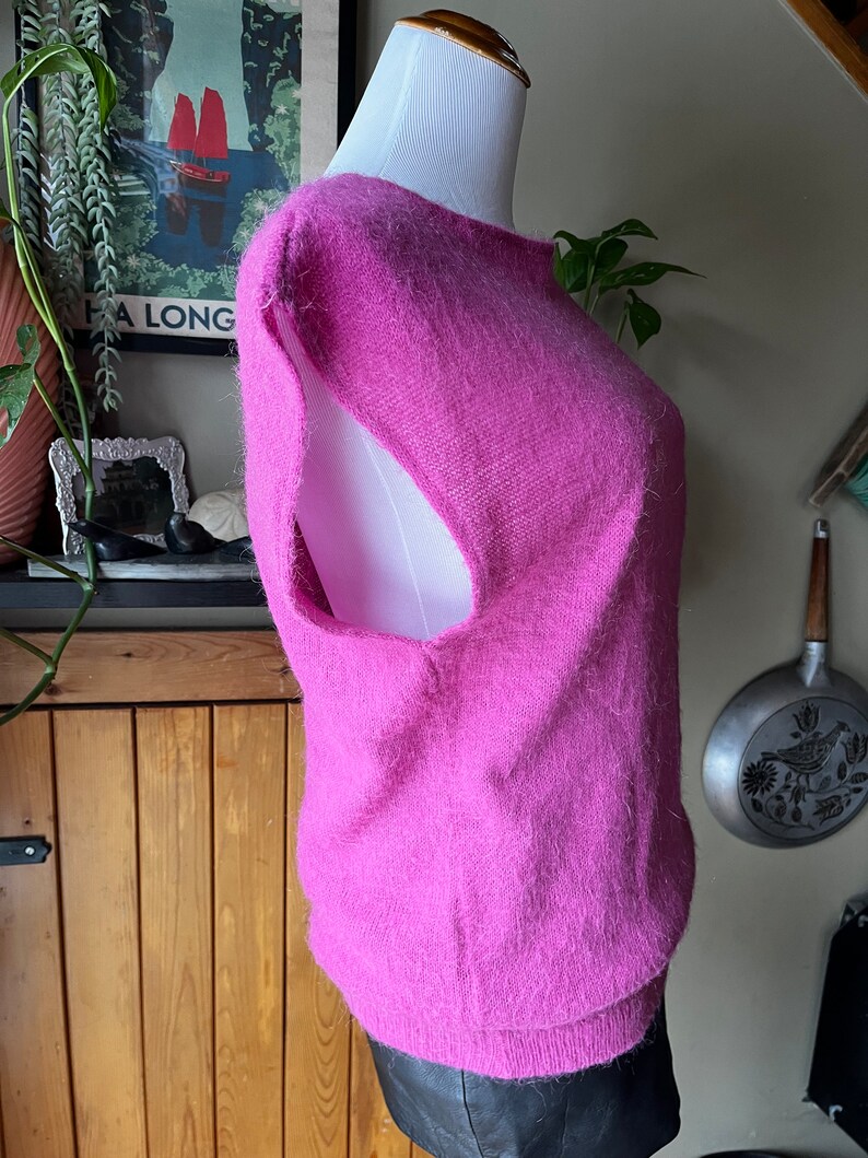 May include: A pink fluffy sleeveless sweater with a scoop neck. The sweater is on a white mannequin and is displayed against a wooden wall.