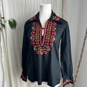 May include: Black long-sleeved blouse featuring a keyhole neckline and embroidered floral pattern in red, yellow, and white. The blouse has a collared neckline and cuffs with matching embroidery. A vintage top.
