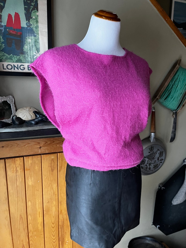 May include: A pink fluffy sleeveless sweater with a round neckline. The sweater is on a mannequin and is paired with a black leather skirt.
