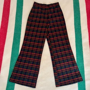 May include: Flared plaid trousers in a traditional tartan design. The trousers have a navy blue background with red and green plaid lines. The garment is displayed on a striped blanket.