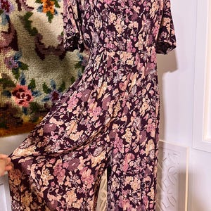 May include: A burgundy floral print jumpsuit with pink and beige flowers. It has a scoop neckline, short sleeves, and wide-leg trousers. The fabric appears soft and flowing, with a detailed floral pattern. The jumpsuit is a one-piece garment.