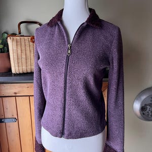 May include: A purple zip-up jacket with a textured knit fabric. The jacket has a dark purple collar and cuffs with a fuzzy texture. A gold-colored zipper runs down the front.
