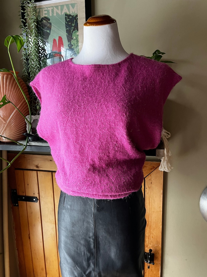 May include: A pink fluffy sleeveless sweater with a round neckline. The sweater is on a white mannequin and is paired with a black leather skirt.