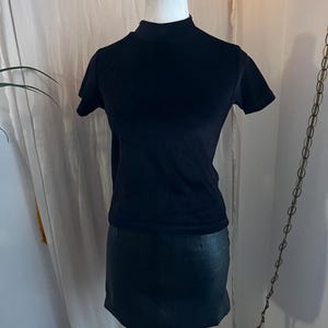 May include: A black short-sleeved top with a mock neck is displayed on a mannequin. Below the top is a black mini skirt. The clothing is displayed against a neutral background.