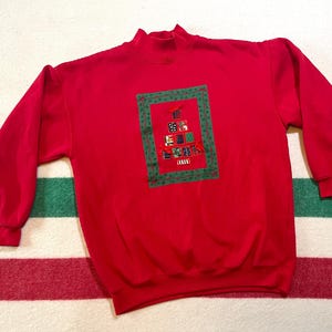 May include: A red sweatshirt with a high neck and long sleeves. It has an embroidered Christmas tree design with gift boxes in various colours, framed by a green border. A festive holiday sweater.