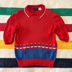 May include: A red knitted top with a white-edged collar and short sleeves. The shirt has a blue and white sailboat pattern at the hem. It is shown on a striped blanket.