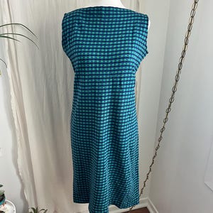 Peut inclure: Robe sans manches, longueur genou, à motif à carreaux bleu sarcelle et bleu marine. La robe est présentée sur un mannequin blanc, mettant en valeur son corsage ajusté et sa jupe droite. Le tissu semble être un tricot texturé.