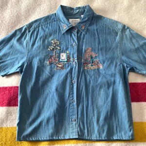 May include: Pale blue denim shirt with short sleeves and embroidered details. The shirt has floral and landscape embroidery on the front. Small embroidered flowers adorn the collar. A casual, everyday top.