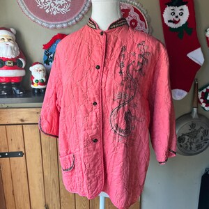 May include: A coral quilted jacket with black button fastenings and a mandarin collar. The jacket has a black embroidered design on the front and black trim on the sleeves and pockets. The jacket is displayed on a mannequin.