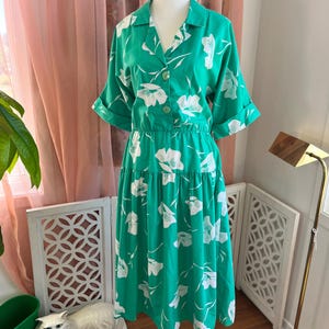 Könnte beinhalten: Ein vintage grünes Kleid mit weißem Blumenmuster. Das Kleid hat einen Hemdkragen, eine Knopfleiste vorne und kurze Ärmel. Die Taille ist gerafft und der Rock fällt bis zu einer Midi-Länge. Das Kleid wird auf einer weißen Schaufensterpuppe präsentiert.