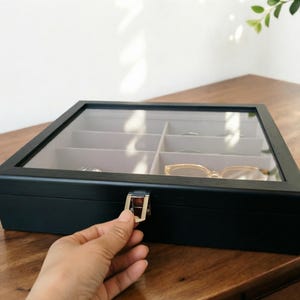 K&ouml;nnte beinhalten: Eine schwarze Brillenbox aus Holz mit einer transparenten Glasoberseite und mehreren F&auml;chern. Die Box ist rechteckig mit einem silbernen Verschluss. Im Inneren befinden sich mehrere Brillen und andere Gegenst&auml;nde. Die Box steht auf einer Holzoberfl&auml;che.