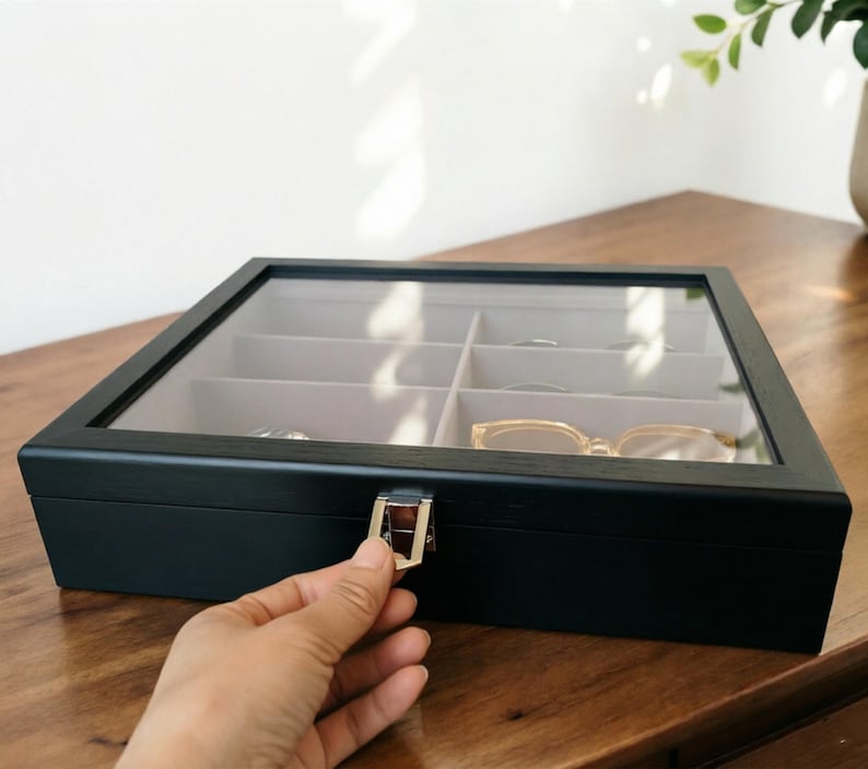 K&ouml;nnte beinhalten: Eine schwarze Brillenbox aus Holz mit einer transparenten Glasoberseite und mehreren F&auml;chern. Die Box ist rechteckig mit einem silbernen Verschluss. Im Inneren befinden sich mehrere Brillen und andere Gegenst&auml;nde. Die Box steht auf einer Holzoberfl&auml;che.