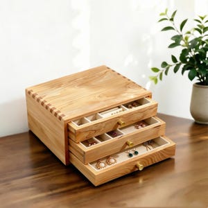 Könnte beinhalten: Ein Schmuckkästchen aus hellem Holz mit drei übereinanderliegenden Schubladen und kleinen Messingknöpfen. Das Innere jeder Schublade ist mit cremefarbenem Stoff ausgekleidet und in mehrere Fächer unterteilt, die verschiedene Ohrringe enthalten.