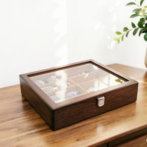 K&ouml;nnte beinhalten: Eine rechteckige, dunkelbraune Brillenbox aus Holz mit einer transparenten Glasoberseite. Die Box ist in sechs F&auml;cher unterteilt, in denen jeweils eine Brille aufbewahrt wird. Ein silberner Verschluss sichert den Deckel. Die Box steht auf einer Holzoberfl&auml;che.