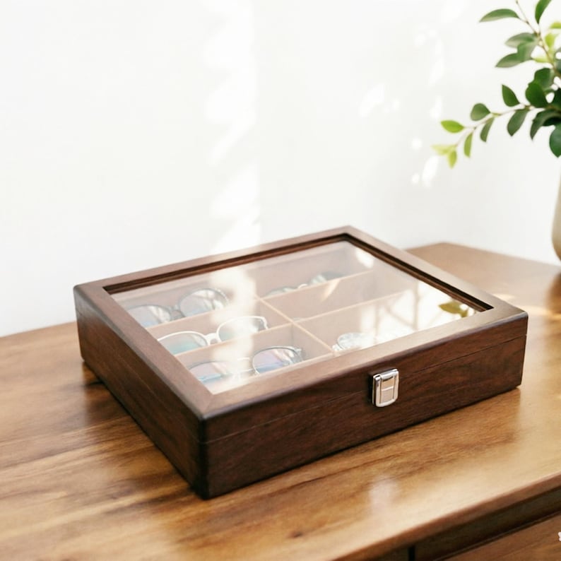 K&ouml;nnte beinhalten: Eine rechteckige, dunkelbraune Brillenbox aus Holz mit einer transparenten Glasoberseite. Die Box ist in sechs F&auml;cher unterteilt, in denen jeweils eine Brille aufbewahrt wird. Ein silberner Verschluss sichert den Deckel. Die Box steht auf einer Holzoberfl&auml;che.