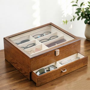 May include: A rectangular wooden storage box with a glass lid and a pull-out drawer. The interior is lined with cream-colored fabric and divided into multiple compartments. Several pairs of eyeglasses are stored inside the box and the drawer.