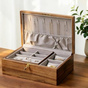 Könnte beinhalten: Geöffnete Holz-Schmuckschatulle mit mehreren Fächern und hellem grauem Innenraum. Die Schatulle ist aus warmtonigem Holz gefertigt und hat ein Schlüsselschloss. Mehrere Halsketten und andere Schmuckstücke sind sichtbar.