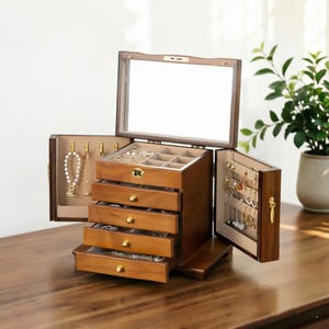 Könnte beinhalten: Eine Holz-Schmuckschatulle mit mehreren Schubladen und Fächern. Die Schatulle hat einen Klappdeckel mit Spiegel und Seitenfächern zum Aufhängen von Schmuck. Die Schatulle ist aus hellbraunem Holz gefertigt und hat goldfarbene Beschläge.