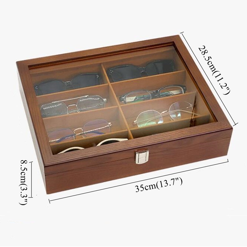 K&ouml;nnte beinhalten: Eine Holz-Brillenaufbewahrungsbox mit transparentem Deckel, die verschiedene Brillen und Sonnenbrillen enth&auml;lt. Die Box ist 35 cm lang, 28,5 cm breit und 8,5 cm hoch. Der Innenraum ist in F&auml;cher unterteilt und hat einen silbernen Verschluss.