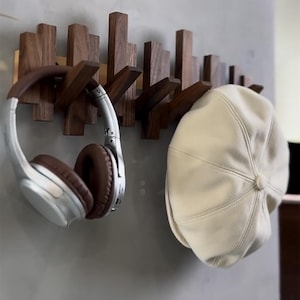 Op de afbeelding: Een houten wandkapstok met meerdere haken. Een bruin-zilveren koptelefoon en een crèmekleurige pet hangen aan de kapstok. De kapstok is gemaakt van donker hout en heeft een modern design.