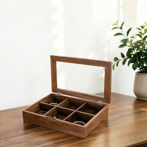 May include: A wooden jewelry box with a clear glass lid, divided into six compartments. The box is open, revealing various bracelets and necklaces. The wood has a warm, brown tone, and the box sits on a wooden surface.