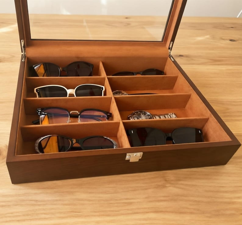 K&ouml;nnte beinhalten: Ein h&ouml;lzerner Sonnenbrillen- und Uhren-Organizer mit Glasdeckel. Der Innenraum ist mit beigem Stoff ausgekleidet und verf&uuml;gt &uuml;ber mehrere F&auml;cher, die verschiedene Sonnenbrillen und eine Uhr aufnehmen. Die Box ist ge&ouml;ffnet und zeigt den Inhalt.