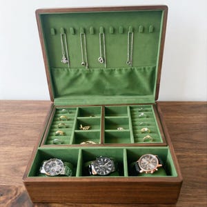 Può includere: Un portagioie in legno con interno in velluto verde. La scatola è aperta, rivelando scomparti per anelli, collane e orologi. Diverse collane d'argento sono appese in alto, mentre anelli e orologi sono ben esposti sotto.