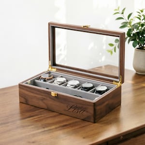 May include: A wooden watch box with a clear glass lid, holding several wristwatches. The box is brown with a gray interior and gold-colored hardware. The name "Lisa" is engraved on the front.