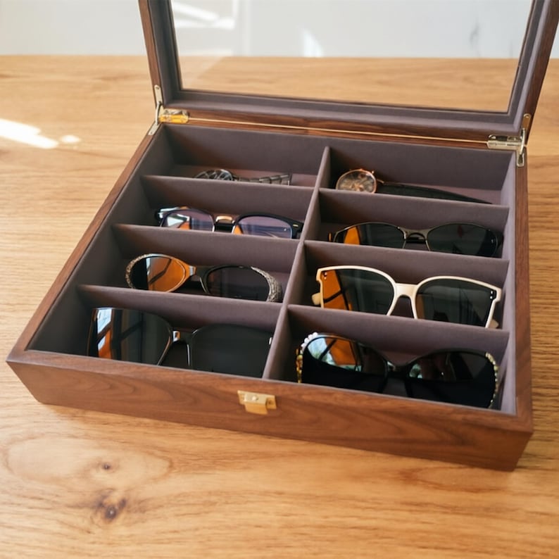 K&ouml;nnte beinhalten: Eine Holz-Sonnenbrillen- und Uhren-Pr&auml;sentationsbox mit Glasdeckel. Der Innenraum ist mit grauem Stoff ausgekleidet und in F&auml;cher unterteilt, die verschiedene Sonnenbrillen und zwei Uhren aufnehmen. Die Box ist ge&ouml;ffnet.
