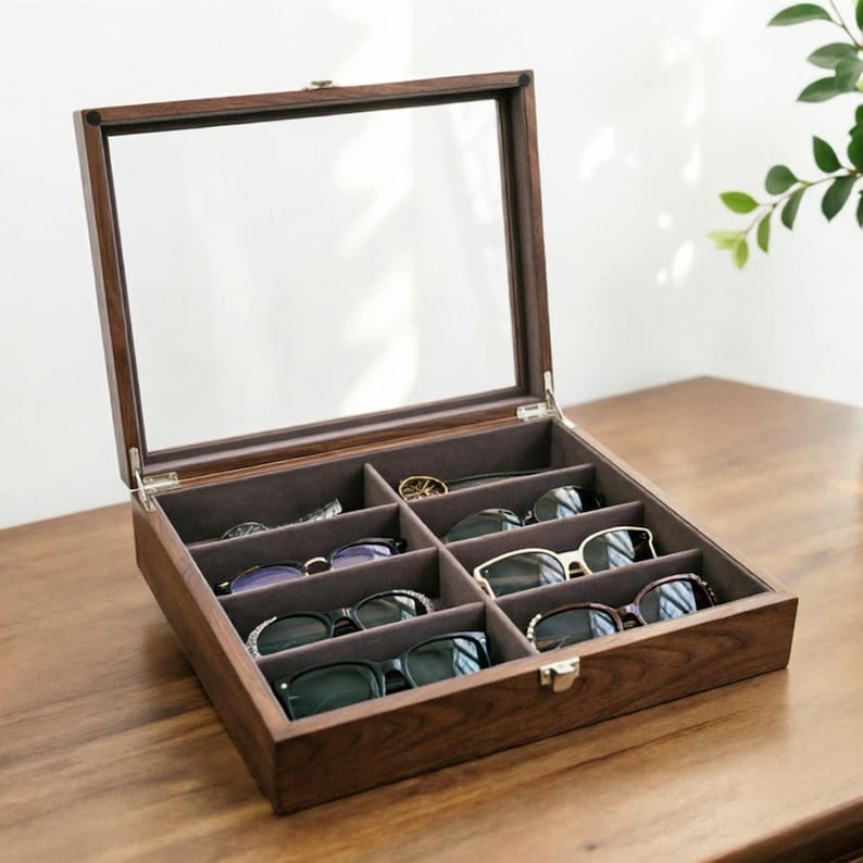 K&ouml;nnte beinhalten: Eine Brillen-Vitrine aus Holz mit klarer Glasoberfl&auml;che, die mehrere Sonnenbrillen und Brillen zeigt. Der Innenraum ist mit einem weichen, grauen Stoff ausgekleidet und das Geh&auml;use ist in F&auml;cher unterteilt. Das Geh&auml;use ist ge&ouml;ffnet und zeigt die Brillen.