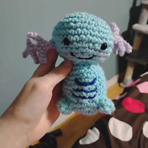 May include: A handmade crochet plush toy in the shape of a cartoon character. The toy is primarily light blue with lavender accents on the side fins. It has black eyes and a stitched smile, with blue detailing on the belly.