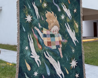 May include: A rectangular woven throw blanket featuring a dark green background with a central figure wearing a multicolored checkered shirt, surrounded by white rabbits and various colorful shooting stars, finished with a fringed edge on all sides.